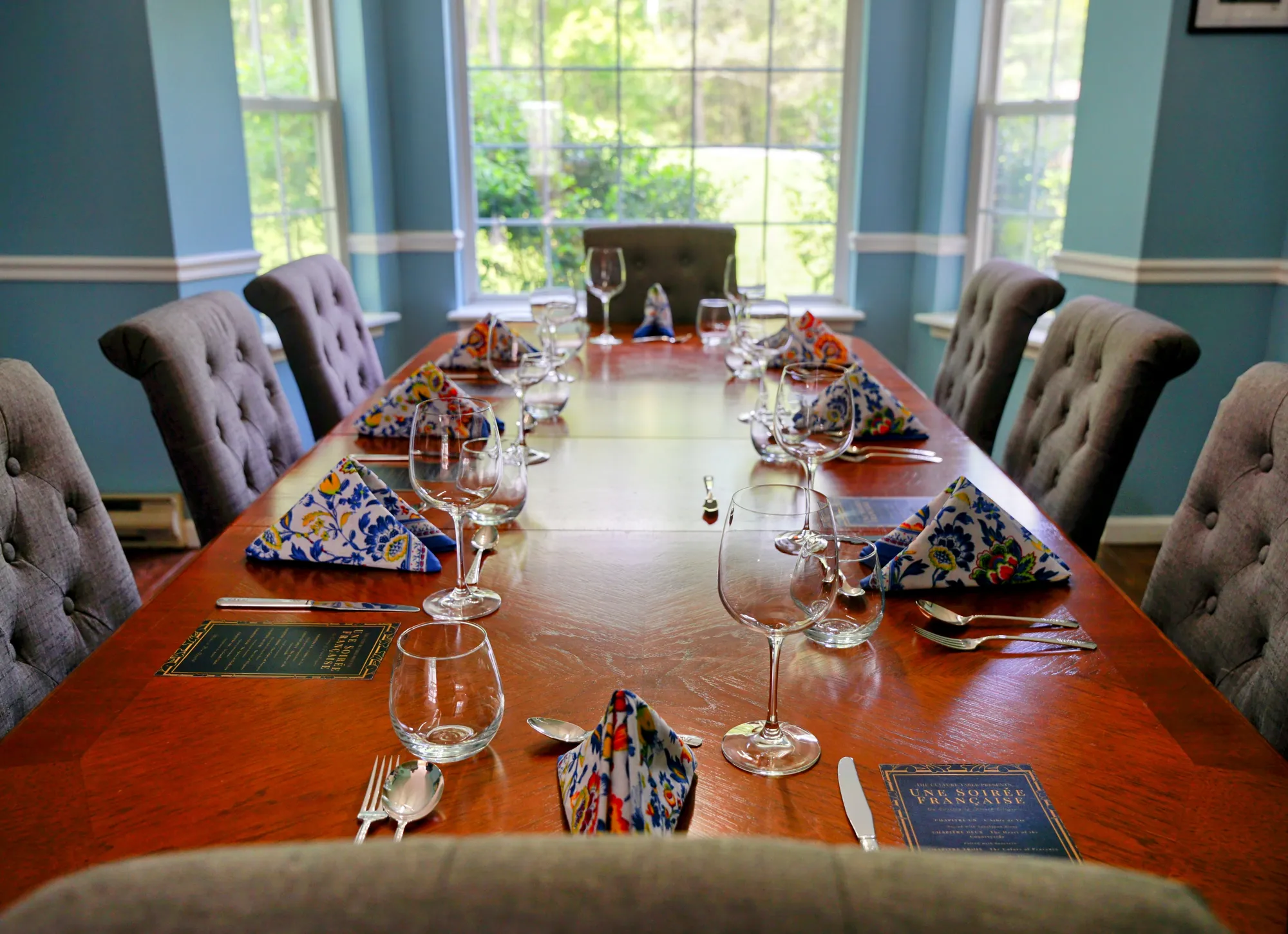 Long candlelit dining table set for ten — wing-backed gray chairs, French menu cards at each setting, blue-and-orange folded napkins, evening light through bay windows.