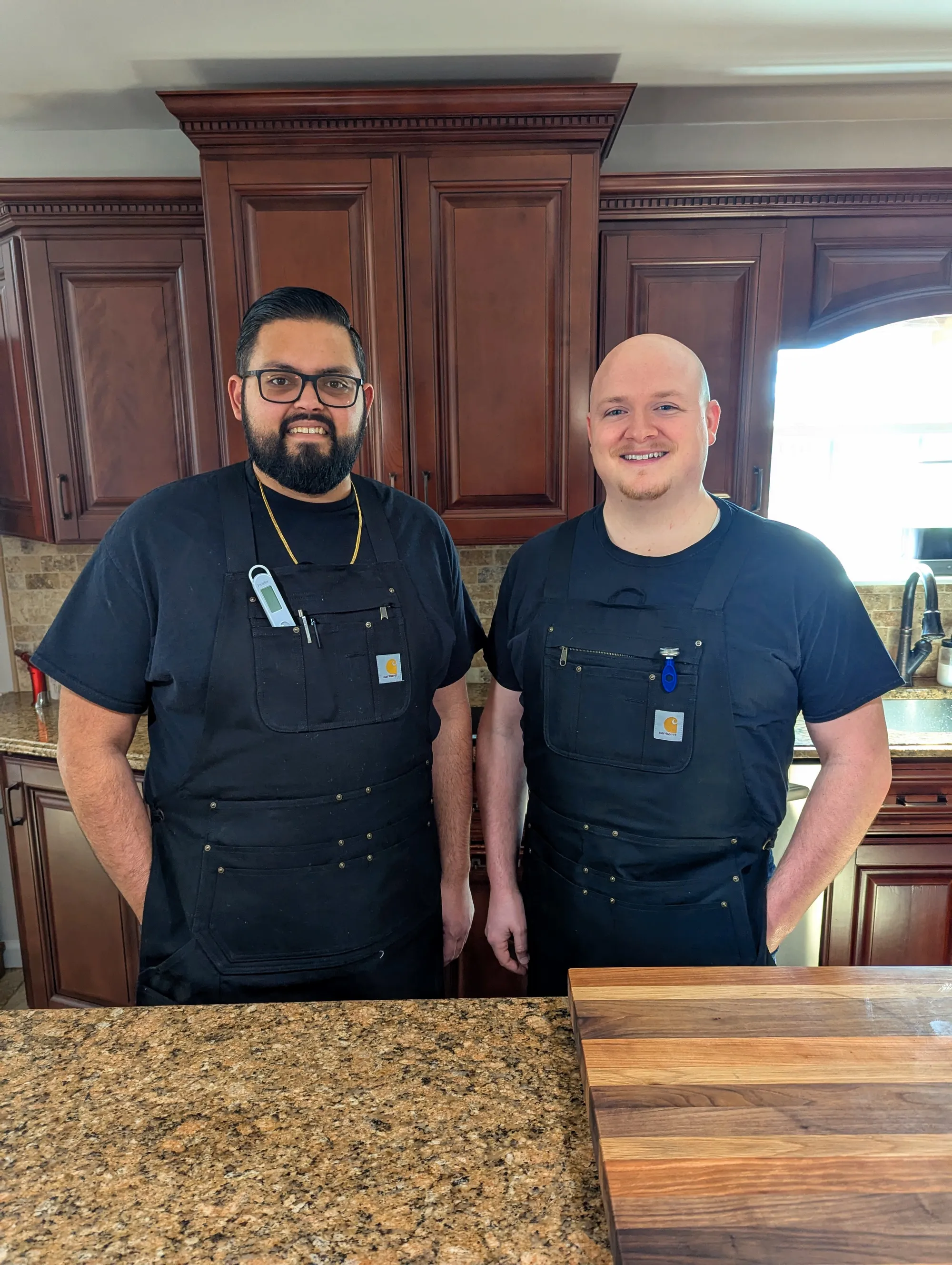 Dylan Lowe and Nalin Dadarwala in matching black work aprons, standing in their kitchen with cherry-wood cabinets behind them.